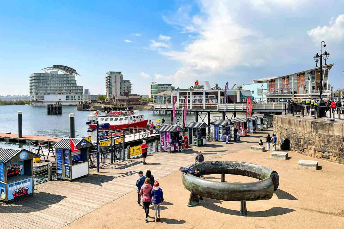 A bustling waterfront promenade with people walking near stalls, a boat in the water, and modern architecture in the distance